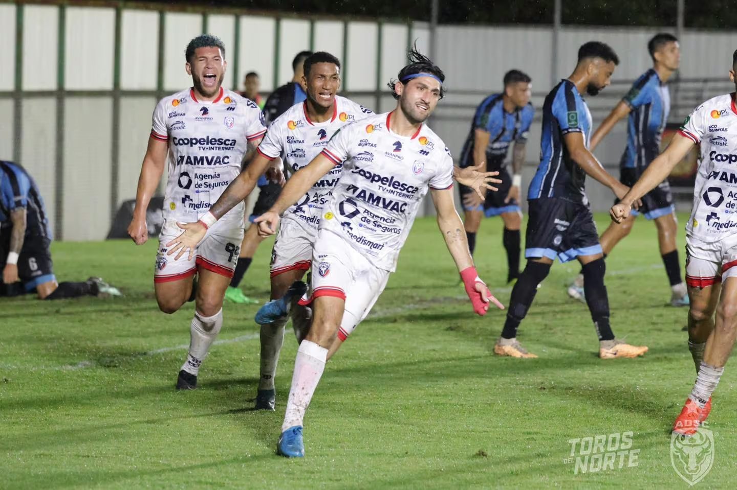 Jugadores de la Asociación Deportiva San Carlos celebran un gol durante un partido de fútbol. Aarón Salazar lidera la celebración con expresión de euforia mientras otros compañeros lo siguen.