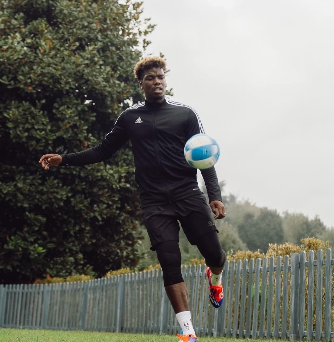 Paul Pogba entrenando al aire libre con ropa deportiva negra, sosteniendo un balón azul y blanco, con un fondo de árboles y una cerca.