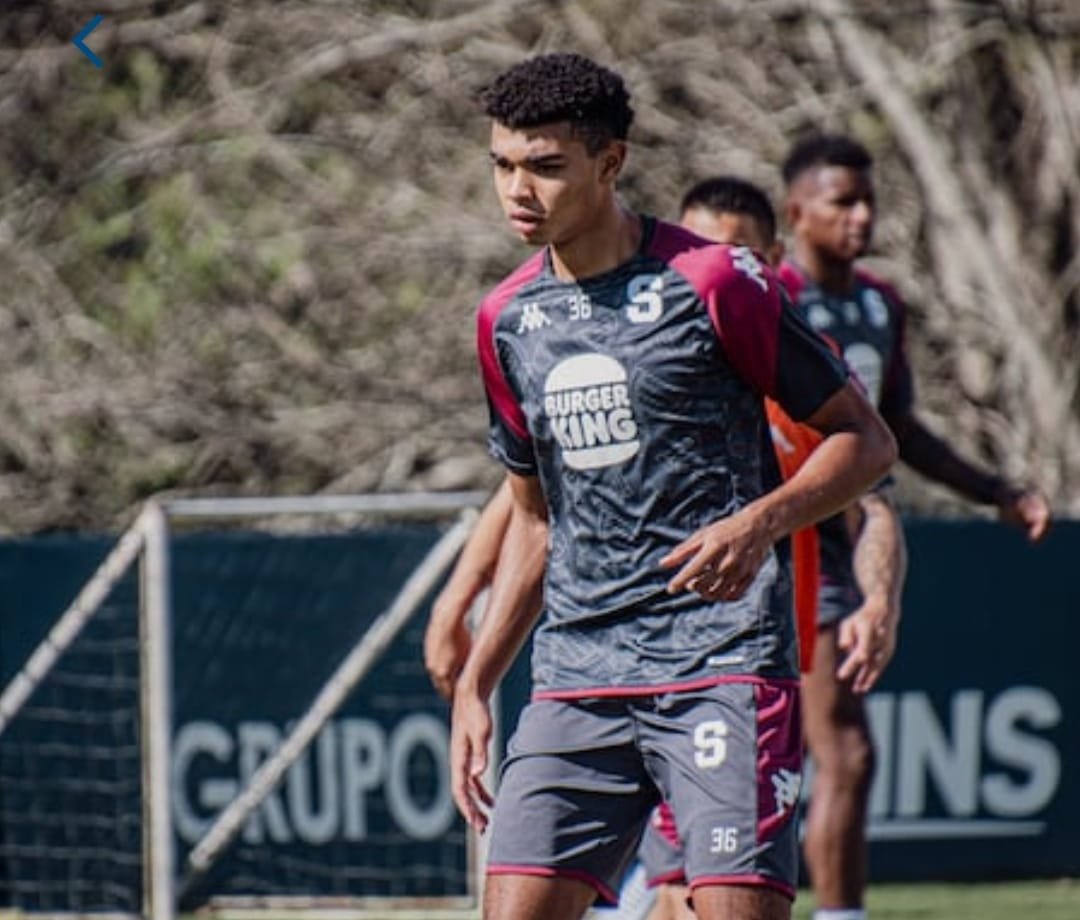 Un jugador del Deportivo Saprissa, vestido con uniforme de entrenamiento negro y burdeos, participa en una sesión de práctica en un campo con redes de fútbol al fondo.